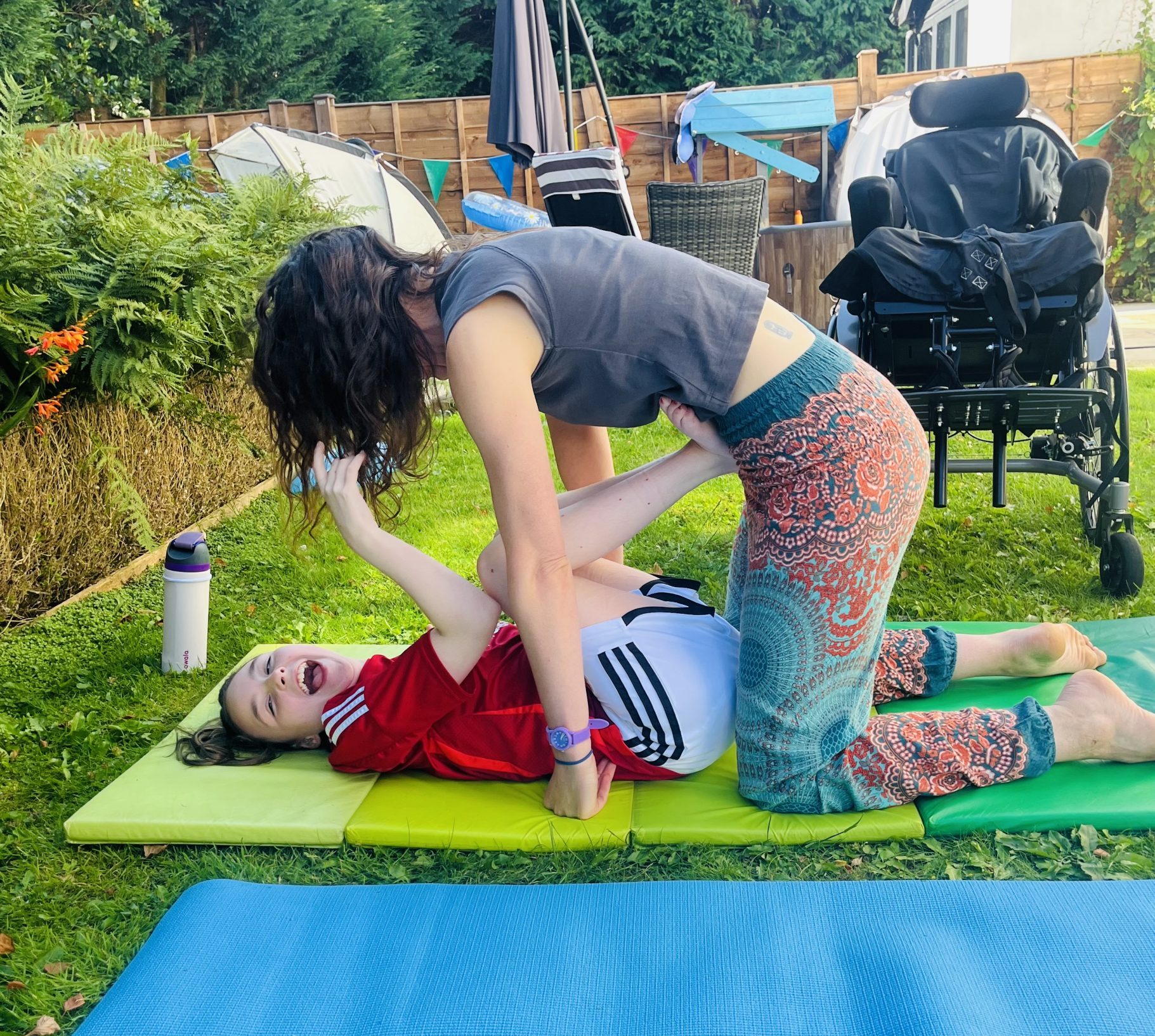 A adult woman working on a yoga mat with a young girl with cerebral palsy, in the garden.
